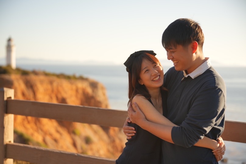 Claire and Paul embracing with smiles overlooking the ocean and lighthouse